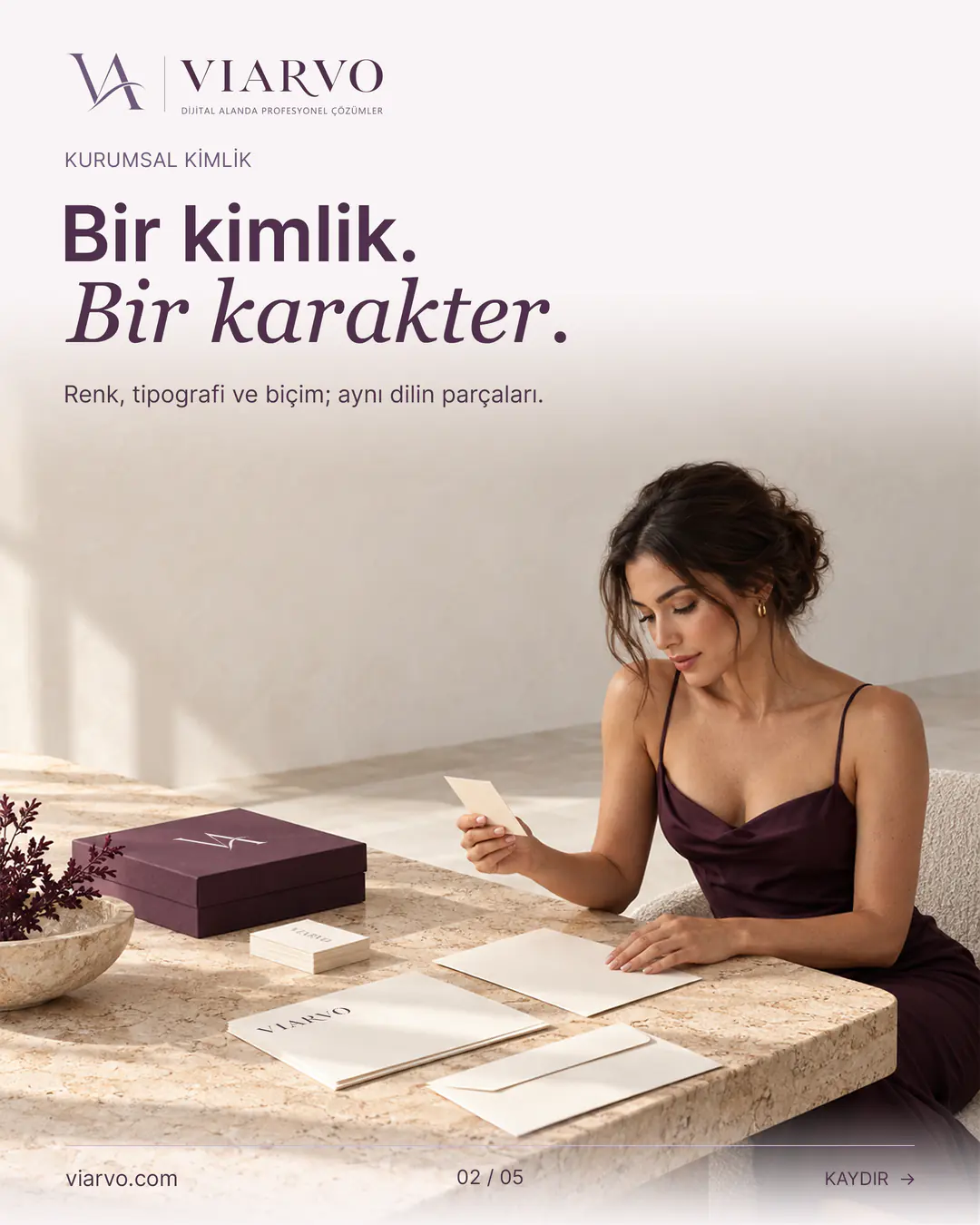 “Bir kimlik. Bir karakter.” başlıklı VIARVO sayfası. Gün ışıklı krem stüdyoda yetişkin kadın tasarımcı, taş masa üzerindeki kartvizit, kâğıt ve mürdüm sunum kutusunu inceliyor. Kurumsal kimlik uygulamaları aynı krem ve mürdüm dili taşıyor.